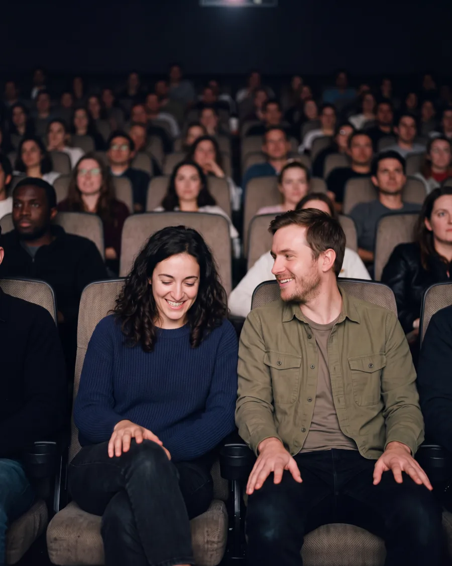 A couple in the front row of a full cinema, she laughs with a hand on her knee while he smiles at her